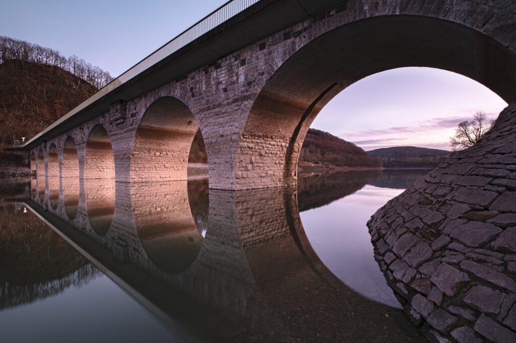Image of a stone bridge with arches over a river, The bridge is reflected in the river surface.